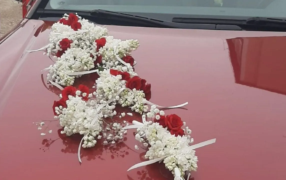Balti ziedi ar sarkanām rozēm un lentēm uz kāzu auto motora pārsega