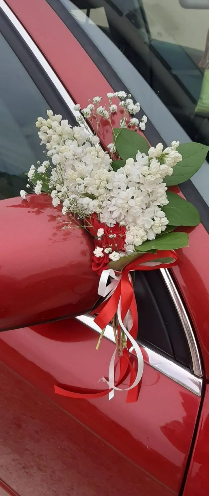 Balts ziedu rotājums ar sarkanām lentēm uz kāzu auto spoguļa